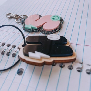 May include: Two keychains with a black and white lightning bolt design. One keychain is pink with glitter and a green smiley face. The other keychain is black with a white smiley face.