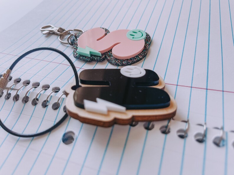 May include: Two keychains with a black and white lightning bolt design. One keychain is pink with glitter and a green smiley face. The other keychain is black with a white smiley face.