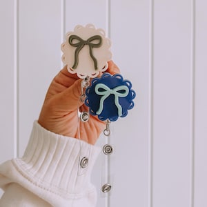 Op de afbeelding: Twee badgehaspels met geschulpte randen en strikaccenten. De ene haspel is wit met een groene strik, de andere is marineblauw met een lichtblauwe strik. Elke haspel heeft een intrekbaar koord en een druksluiting.