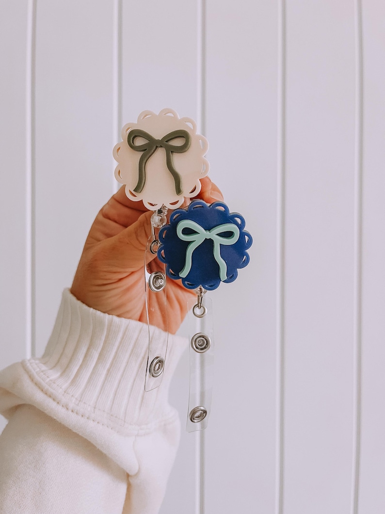 Op de afbeelding: Twee badgehaspels met geschulpte randen en strikaccenten. De ene haspel is wit met een groene strik, de andere is marineblauw met een lichtblauwe strik. Elke haspel heeft een intrekbaar koord en een druksluiting.