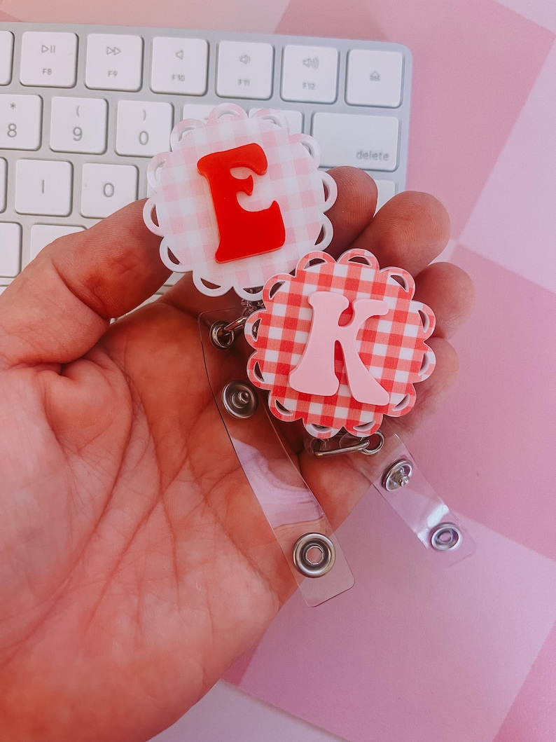 K&ouml;nnte beinhalten: Zwei runde Ausweishalter mit gewelltem Rand und einem rosa-wei&szlig;en Vichy-Muster. Ein Halter zeigt den roten Buchstaben E, der andere den hellrosa Buchstaben K. Beide verf&uuml;gen &uuml;ber transparente Kunststoffb&auml;nder mit silbernen Druckkn&ouml;pfen.