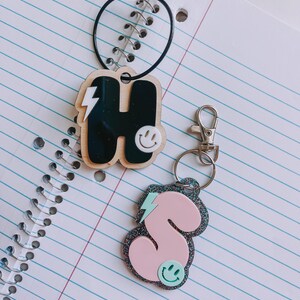 May include: Two keychains with black and pink letter charms. The charms are shaped like the letters "H" and "S" and have a lightning bolt and smiley face design.
