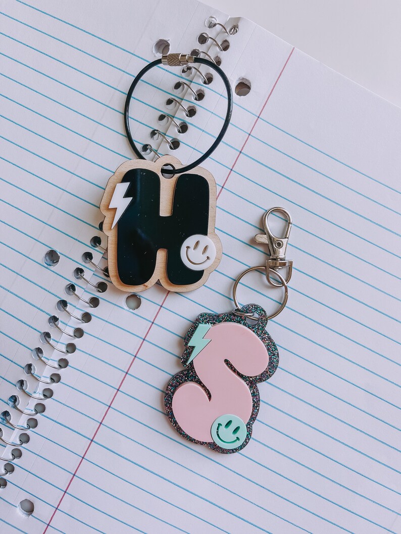 May include: Two keychains with black and pink letter charms. The charms are shaped like the letters "H" and "S" and have a lightning bolt and smiley face design.