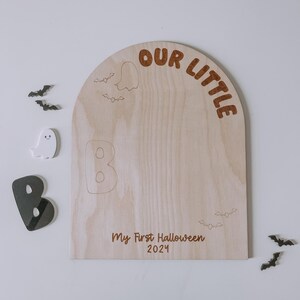 May include: A wooden sign shaped like an arch with the text "Our Little" at the top. The sign is decorated with bats and ghosts. The text "My First Halloween 2021" is written below the arch.