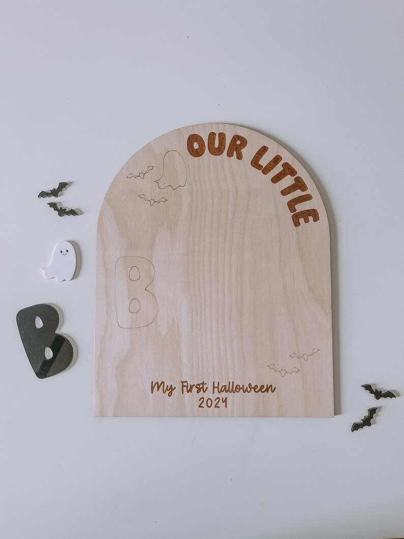 May include: A wooden sign shaped like an arch with the text "Our Little" at the top. The sign is decorated with bats and ghosts. The text "My First Halloween 2021" is written below the arch.