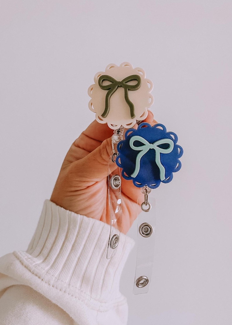 Op de afbeelding: Twee badgehaspels met geschulpte randen en strikdesigns. E&eacute;n haspel is lichtbeige met een donkergroene strik, de andere is donkerblauw met een lichtblauwe strik. Beide hebben transparante, intrekbare plastic koorden en metalen clips.