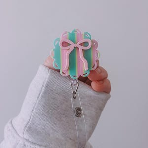 May include: A pastel-colored badge reel with a scalloped edge and a pink bow design. The badge reel has a clear plastic strap with a metal snap closure. The background is a solid light gray.