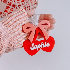 May include: A red and pink cherry-shaped ornament with a white ribbon and the name "Sophie" written in white letters.