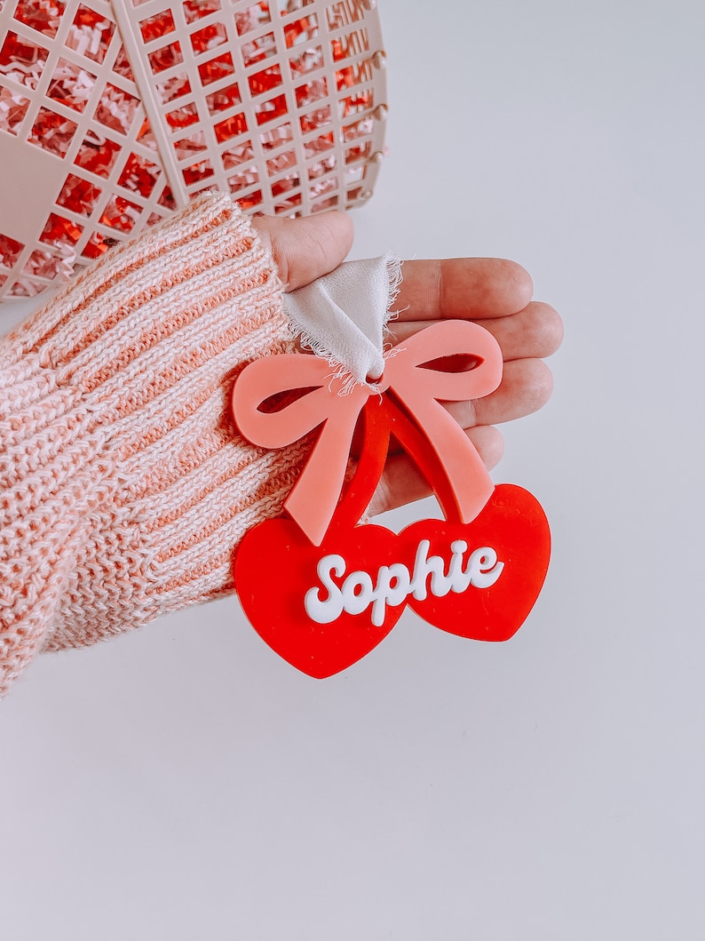 May include: A red and pink cherry-shaped ornament with a white ribbon and the name "Sophie" written in white letters.