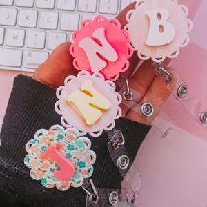 K&ouml;nnte beinhalten: Drei runde Ausweishalter mit gewelltem Rand und erhabenen Buchstaben. Ein Halter ist leuchtend pink mit einem N, einer ist blassrosa gestreift mit einem B und einer hat ein Blumenmuster mit einem pinken J. Jeder verf&uuml;gt &uuml;ber ein transparentes Band.