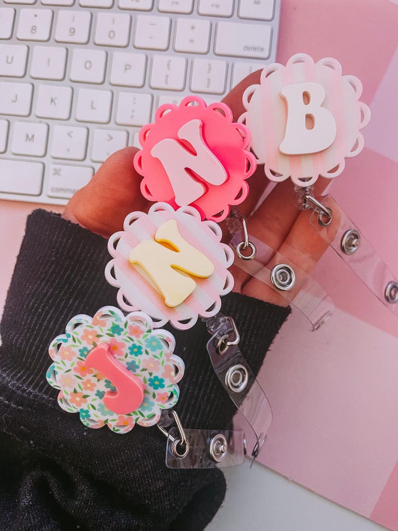 K&ouml;nnte beinhalten: Drei runde Ausweishalter mit gewelltem Rand und erhabenen Buchstaben. Ein Halter ist leuchtend pink mit einem N, einer ist blassrosa gestreift mit einem B und einer hat ein Blumenmuster mit einem pinken J. Jeder verf&uuml;gt &uuml;ber ein transparentes Band.