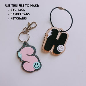 May include: Two keychains with letter charms. The keychain on the left is pink with a glitter finish and a lightning bolt design. The keychain on the right is black with a lightning bolt design. Both charms have a smiley face.