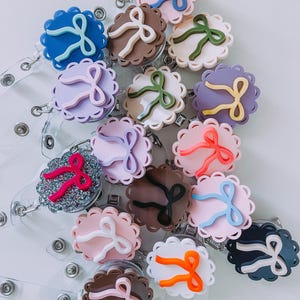 Op de afbeelding: Een verzameling badge reels met een geschulpte rand en een strikaccent. De reels zijn er in verschillende kleuren, waaronder blauw, roze, bruin en paars. Elke reel heeft een doorzichtige plastic band en een metalen clip.