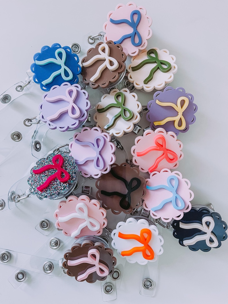 Op de afbeelding: Een verzameling badge reels met een geschulpte rand en een strikaccent. De reels zijn er in verschillende kleuren, waaronder blauw, roze, bruin en paars. Elke reel heeft een doorzichtige plastic band en een metalen clip.