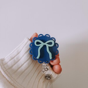 Op de afbeelding: Een blauwe badge reel met een geschulpte rand en een lichtblauw strikdesign. De badge reel wordt vastgehouden door een persoon die een witte trui draagt.