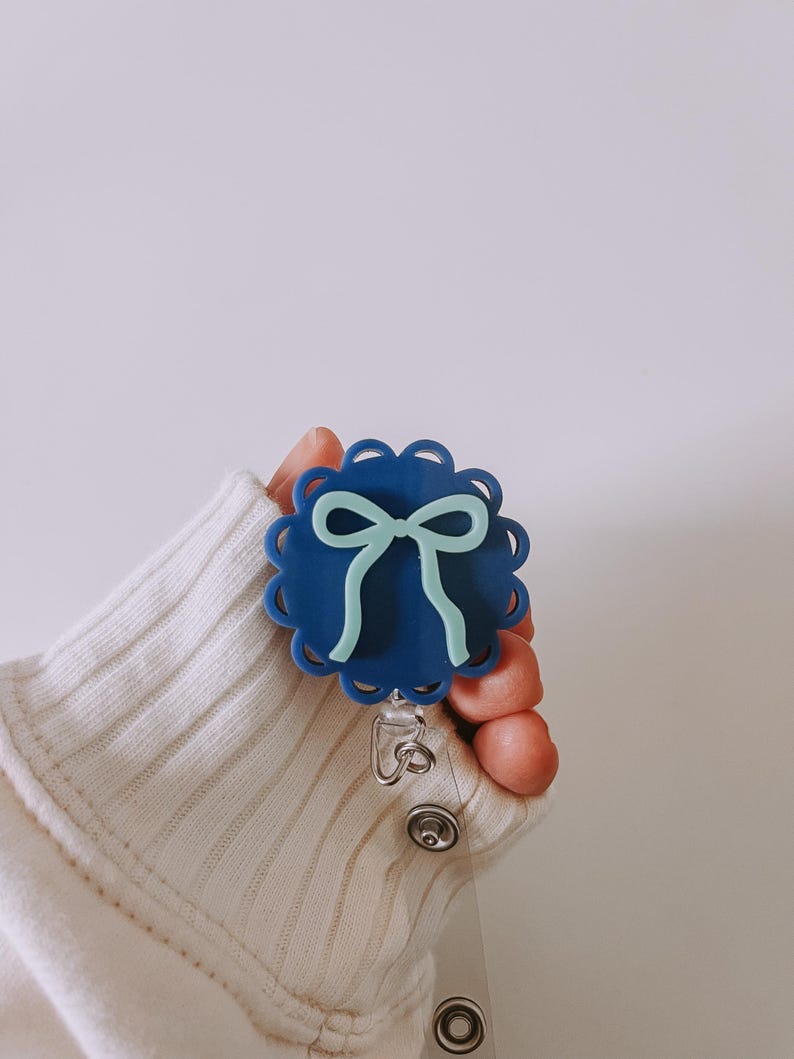Op de afbeelding: Een blauwe badge reel met een geschulpte rand en een lichtblauw strikdesign. De badge reel wordt vastgehouden door een persoon die een witte trui draagt.