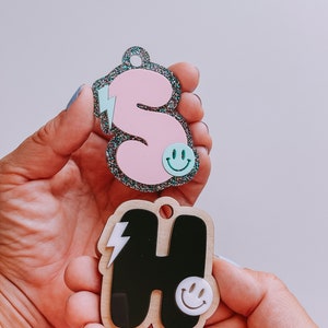 May include: Two wooden letter charms, one pink with glitter and the letter "S" and the other black with the letter "H". Both charms have a lightning bolt and a smiley face.