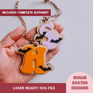 May include: Two wooden Halloween-themed ornaments, one orange letter "H" with bats and one pink letter "S" with bats. The ornaments are hanging from a string and are held in a hand. The text "INCLUDES COMPLETE ALPHABET" is at the top of the image and "LASER READY! SVG FILE" is at the bottom of the image. The text "SUGAR KOATED DESIGNS" is in a circle at the bottom right of the image.
