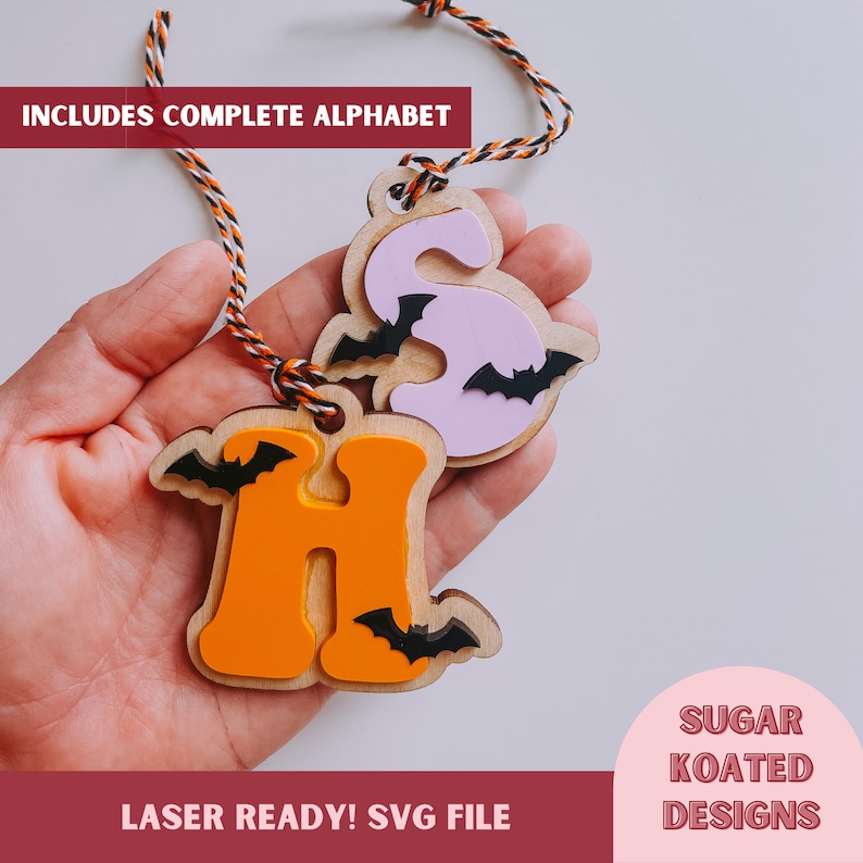 May include: Two wooden Halloween-themed ornaments, one orange letter "H" with bats and one pink letter "S" with bats. The ornaments are hanging from a string and are held in a hand. The text "INCLUDES COMPLETE ALPHABET" is at the top of the image and "LASER READY! SVG FILE" is at the bottom of the image. The text "SUGAR KOATED DESIGNS" is in a circle at the bottom right of the image.