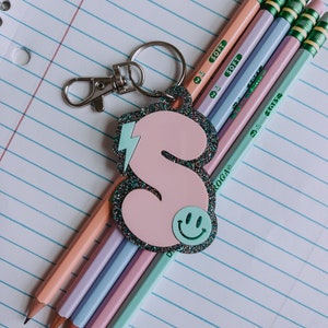 May include: A pink glitter keychain with a lightning bolt and a smiley face. The keychain is attached to a silver key ring and is hanging from a stack of five pastel colored pencils with the word "SOFT" printed on each pencil.