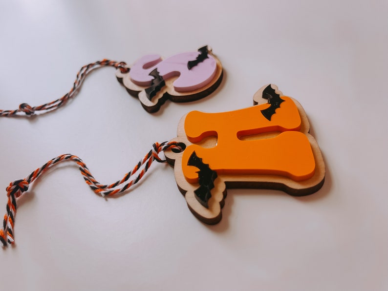 May include: Two wooden Halloween ornaments shaped like the letters "H" and "O" with bats attached. The "H" is orange and the "O" is purple. Both ornaments have twine attached for hanging.