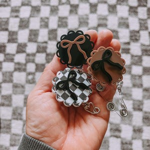 Op de afbeelding: Drie badgehaspels met geschulpte randen en strikaccenten. E&eacute;n is zwart met een bruine strik, een andere is bruin met een zwarte strik, en de derde heeft een zwart-wit geblokt patroon met een zwarte strik. Elke haspel heeft een doorzichtig, intrekbaar plastic koord.