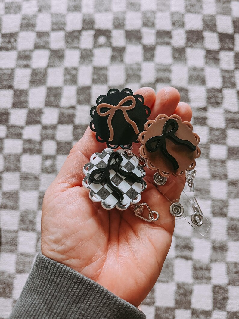 Op de afbeelding: Drie badgehaspels met geschulpte randen en strikaccenten. E&eacute;n is zwart met een bruine strik, een andere is bruin met een zwarte strik, en de derde heeft een zwart-wit geblokt patroon met een zwarte strik. Elke haspel heeft een doorzichtig, intrekbaar plastic koord.