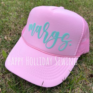 May include: Pink trucker hat with a turquoise blue cursive name "Margs" embroidered on the front. The hat has a mesh back and a pink brim. The text "HAPPY HOLIDAY SEWING" is printed on the brim.