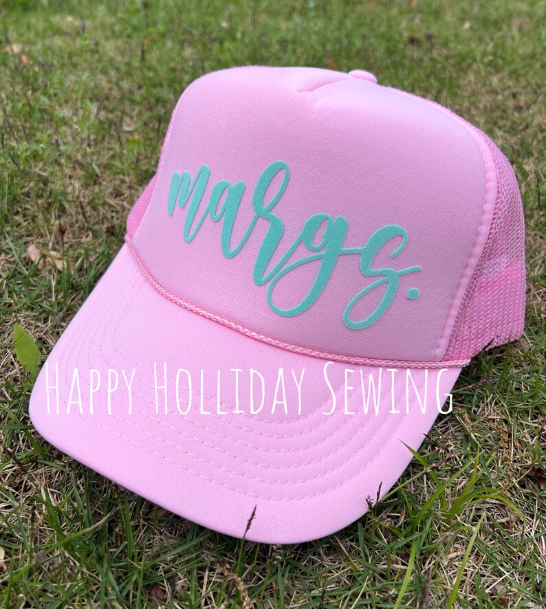 May include: Pink trucker hat with a turquoise blue cursive name "Margs" embroidered on the front. The hat has a mesh back and a pink brim. The text "HAPPY HOLIDAY SEWING" is printed on the brim.