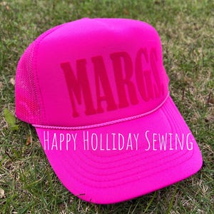 May include: Pink trucker hat with the name "MARG" embroidered in pink on the front. The hat has a black mesh back and a white embroidered label that reads "HAPPY HOLIDAY SEWING".