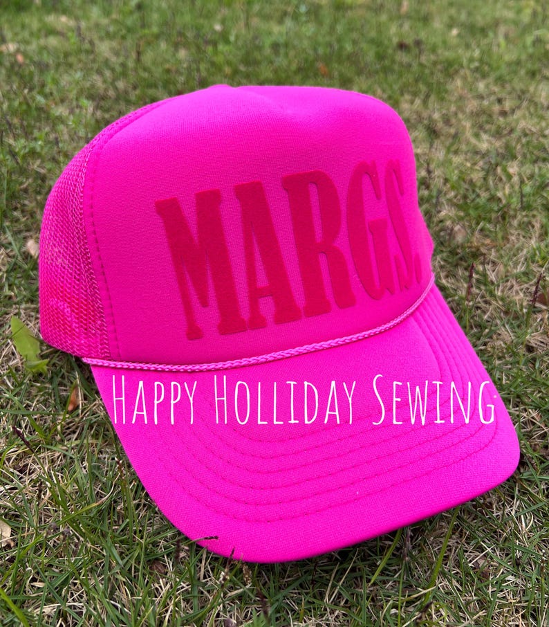 May include: Pink trucker hat with the name "MARG" embroidered in pink on the front. The hat has a black mesh back and a white embroidered label that reads "HAPPY HOLIDAY SEWING".