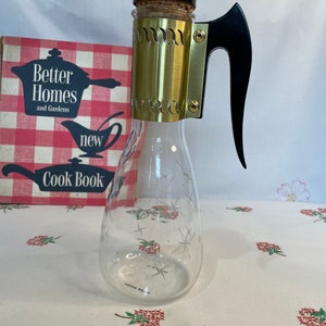May include: Vintage glass cruet with a black handle and stopper, accented with a gold-toned metal band. The clear cruet features etched starburst designs. A vintage cookbook is visible in the background.