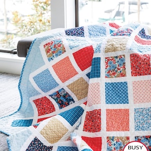May include: A patchwork quilt featuring a grid of square fabric blocks in various floral and geometric patterns in shades of red, blue, and tan, separated by white sashing. The quilt has a visible quilted texture and a Busy Hands Quilts logo.