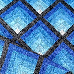 May include: A blue and black quilt with a geometric pattern. The quilt is made of fabric with a swirl design. The quilt is folded in half and the edge is visible. The quilt is on a white surface. The text "BUSY HANDS QUILTS" is visible in the lower right corner of the image.