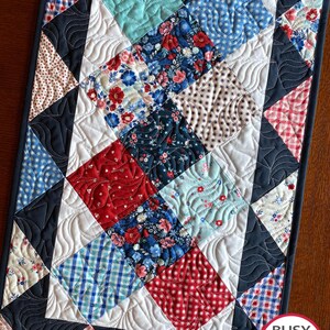 Peut inclure: Chemin de table matelassé avec un motif patchwork. Il présente des carrés de différentes couleurs et motifs, notamment des imprimés floraux et vichy rouges, bleus et blancs. Les bords sont bordés de tissu bleu foncé et la couture a un motif ondulé.