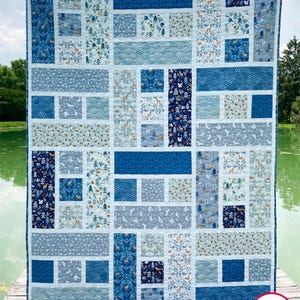 May include: A handmade quilt with a patchwork design in shades of blue, white, and gray. The quilt features various patterns, including forest scenes with deer and trees. The quilt is displayed outdoors, with the text "Busy Hands Quilts" visible.