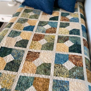 Pode incluir: Uma colcha com um padrão geométrico em tons de marrom, verde e azul. A colcha é feita com uma variedade de tecidos e tem um design acolchoado. A colcha está sobre uma cama com uma cabeceira branca e dois travesseiros azuis. A colcha é feita à mão e tem uma etiqueta que diz "Busy Hands Quilts".