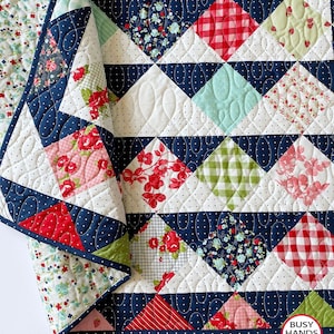 May include: A quilt with a navy blue background and a patchwork design featuring red, green, pink, and white fabrics. The quilt is quilted with a swirl pattern. The quilt is folded in half and the edge of the quilt is visible. The quilt is labeled "Busy Hands Quilts".