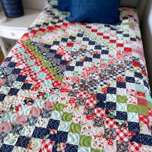 Op de afbeelding: Een patchworkdeken met verschillende bloemen- en ruitpatronen in tinten rood, roze, blauw, groen en wit. De deken is gestikt met een golvend patroon. De deken is gelabeld met "Busy Hands Quilts".