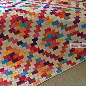 Puede incluir: Una colcha colorida con un patrón geométrico de cuadrados y rectángulos en tonos de rojo, naranja, amarillo, verde, azul y morado. La colcha está acolchada con un patrón de espiral y tiene un fondo blanco. El texto "Busy Hands Quilts" es visible en la esquina inferior derecha de la imagen.