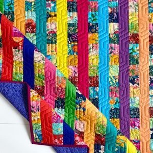 May include: A colorful quilt with a rainbow pattern and a geometric design. The quilt is made of various fabrics with floral prints. The quilt is folded in half and the back side is a blue fabric with white polka dots. The quilt has a label that says "Busy Hands Quilts" and "Buy Handmade".