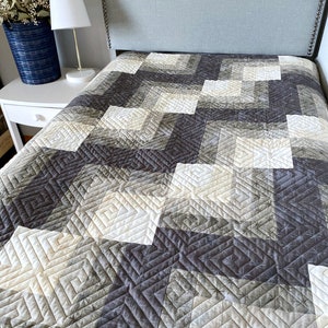 Op de afbeelding: Een grijs en wit quilt met een geometrisch patroon. De quilt is gemaakt van verschillende stoffen en heeft een subtiel ombre-effect. De quilt is te zien op een bed met een grijs hoofdeinde. De quilt is gelabeld met "Busy Hands Quilts".