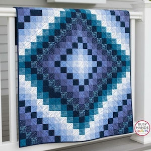 May include: A square quilt featuring a gradient pattern of small fabric squares in shades of navy, royal blue, teal, and white. The design radiates from a light center to dark edges. A logo in the corner reads BUSY HANDS QUILTS.
