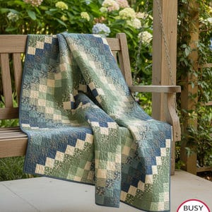 May include: A patchwork quilt featuring a gradient of blue, green, and cream fabric squares arranged in a diagonal pattern. The quilt displays intricate machine quilting stitches across the surface and is finished with a solid blue fabric binding.