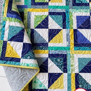 Puede incluir: Una colcha con un patrón geométrico en tonos de azul, verde, amarillo y blanco. La colcha está hecha de cuadrados y triángulos, y la tela tiene una variedad de patrones. La colcha está doblada por la mitad, y el borde de la colcha es visible. La colcha está sobre una superficie blanca. El texto "BUSY HANDS QUILTS" es visible en la esquina inferior derecha de la imagen.