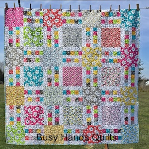 May include: A colorful quilt with a patchwork design featuring squares of different floral patterns and colors. The quilt is quilted with a circular pattern and has a white border. The quilt is hanging on a clothesline with clothespins. The text "Busy Hands Quilts" is printed at the bottom of the quilt.