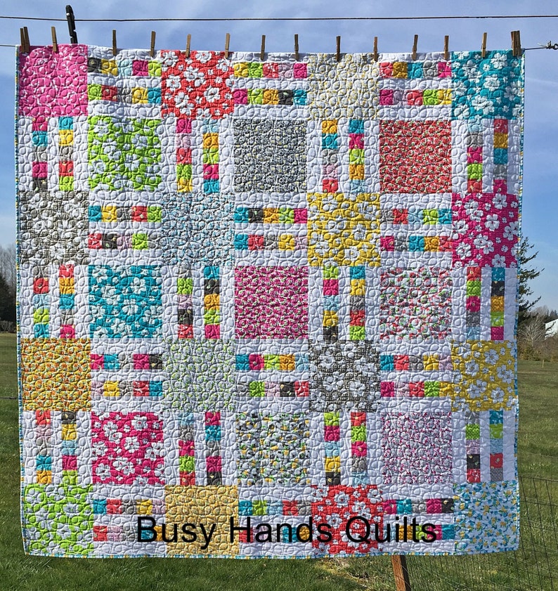 May include: A colorful quilt with a patchwork design featuring squares of different floral patterns and colors. The quilt is quilted with a circular pattern and has a white border. The quilt is hanging on a clothesline with clothespins. The text "Busy Hands Quilts" is printed at the bottom of the quilt.