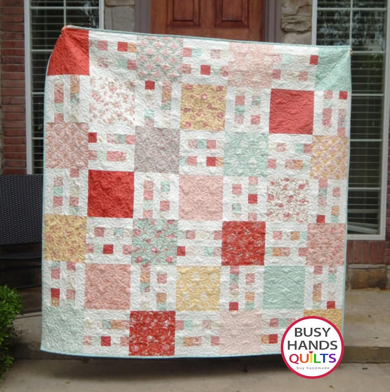 May include: A patchwork quilt with a variety of floral patterns in shades of pink, red, yellow, and green. The quilt is made up of squares and rectangles, and it is quilted with a white thread. The quilt is hanging on a clothesline in front of a brick wall. The quilt is labeled "Busy Hands Quilts" in the bottom right corner.