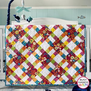 May include: A colorful baby quilt with a criss-cross pattern and a white background. The quilt is made with floral fabric in shades of red, yellow, green, and blue. The quilt is quilted with a circular pattern. The quilt is made by Busy Hands Quilts.