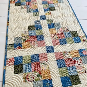 May include: A white quilted table runner with a patchwork design featuring blue, green, red, and yellow squares. The quilting pattern is a series of curved lines. The table runner has the text "Busy Hands Quilts" in the bottom right corner.